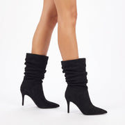 Black slouchy suede ankle boots with pointed toes and kitten heels, shown on a model's feet against a white studio background.